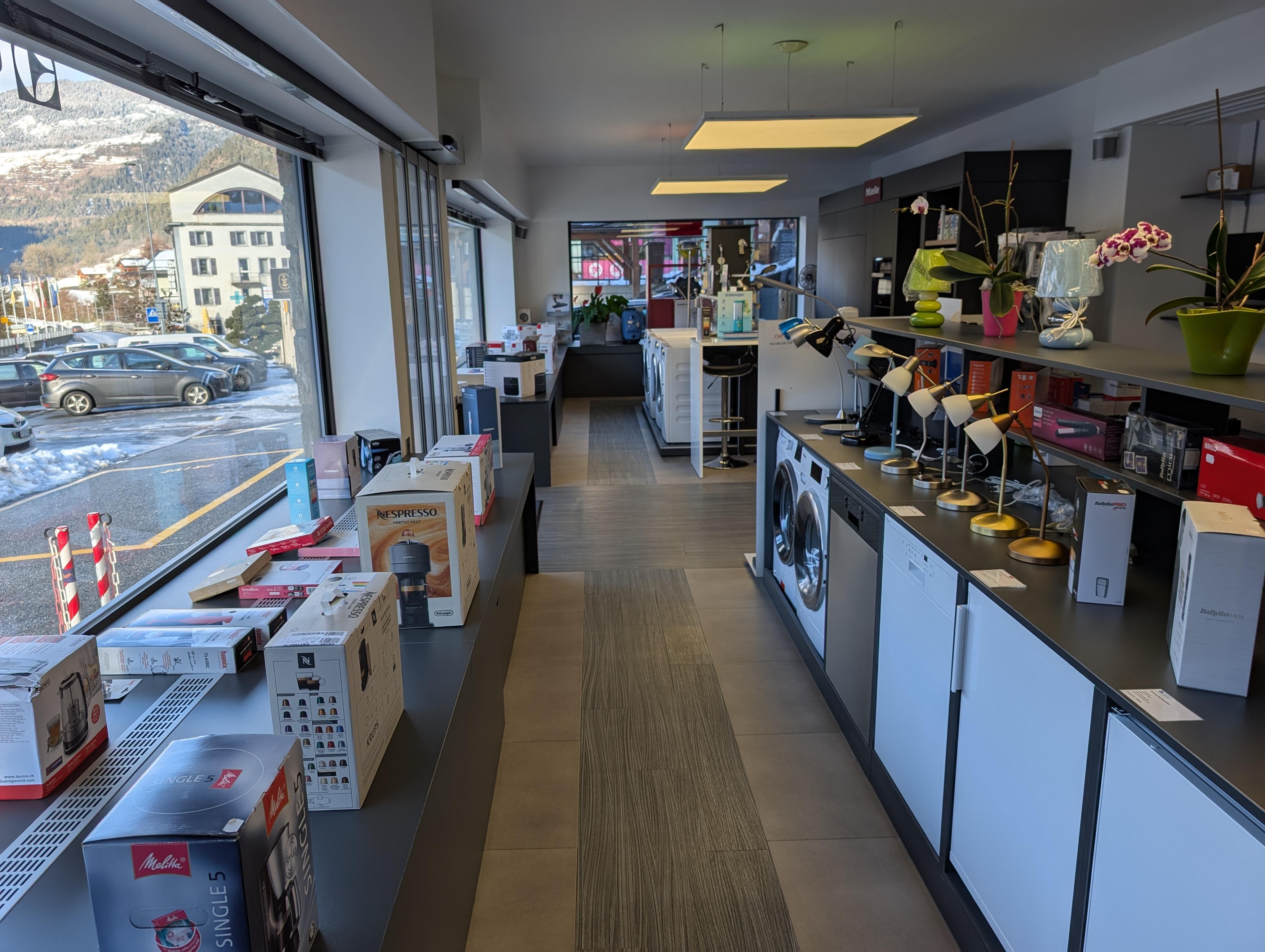 Espace showroom moderne et chaleureux à Sion présentant des machines à café Nespresso et des appareils ménagers Miele sur des étagères en bois clair, éclairage doux